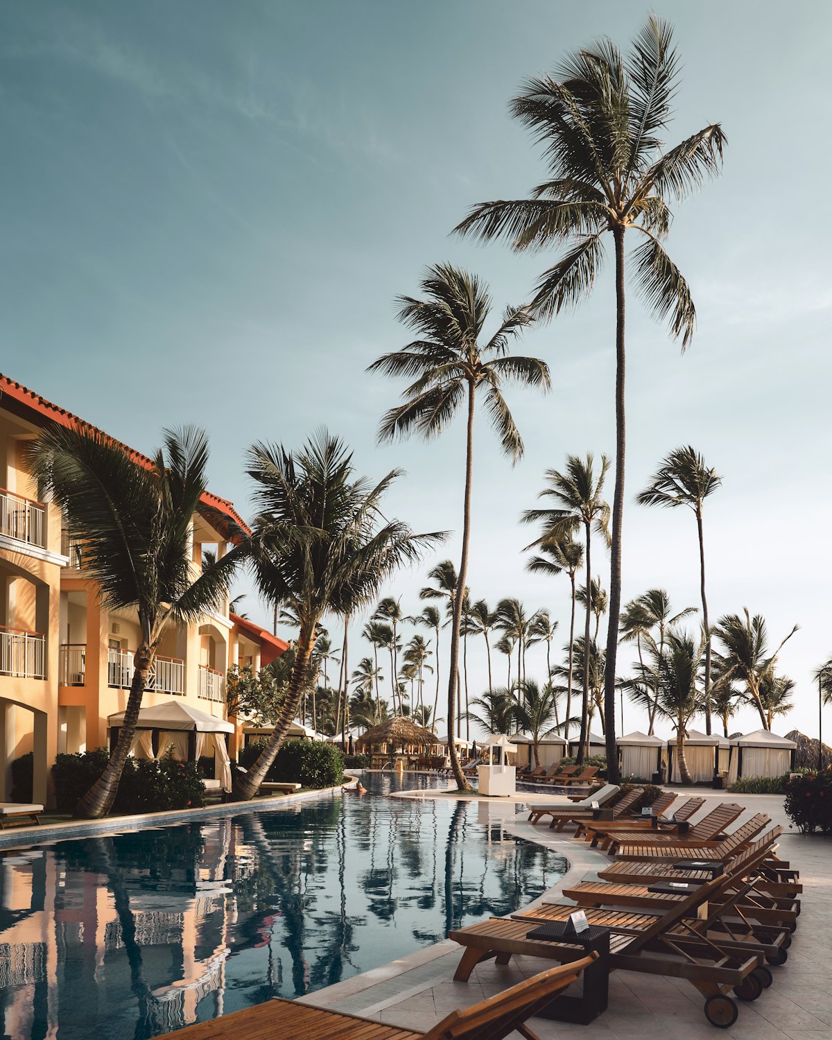 Resort pool lined with palms, cabanas and timber loungers in morning light